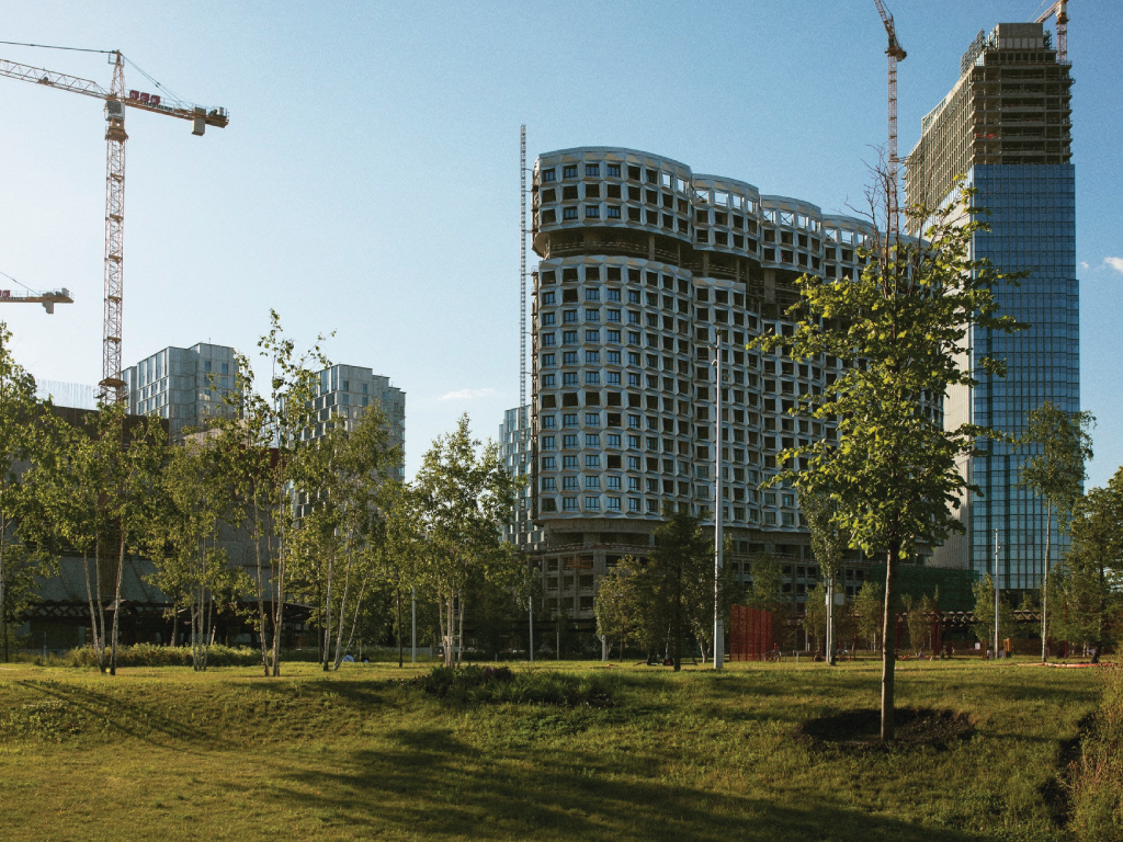 Modern buildings under construction rise behind a green, grassy area with young trees.